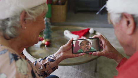 pareja caucásica mayor sonriente usando un teléfono inteligente para una videollamada de navidad con un niño en la pantalla