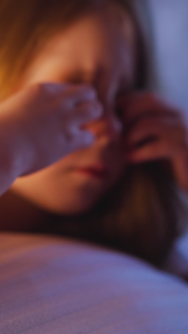 Upset little daughter with loose hair wipes away tears holding large white pillow on bed in partially illuminated bedroom at moon light in evening slow motion