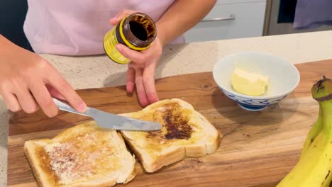 person spreads butter and dark yeast extract on toasted bread slices in a brightly lit kitchen, using a knife and wooden cutting board, with a banana nearby