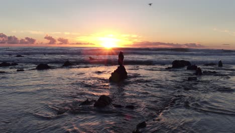 stunning view of the sunset at low tide in laguna beach with a drone flying and surfers surfing in the background as the sun peaks out amongst rocks and reflects off the water