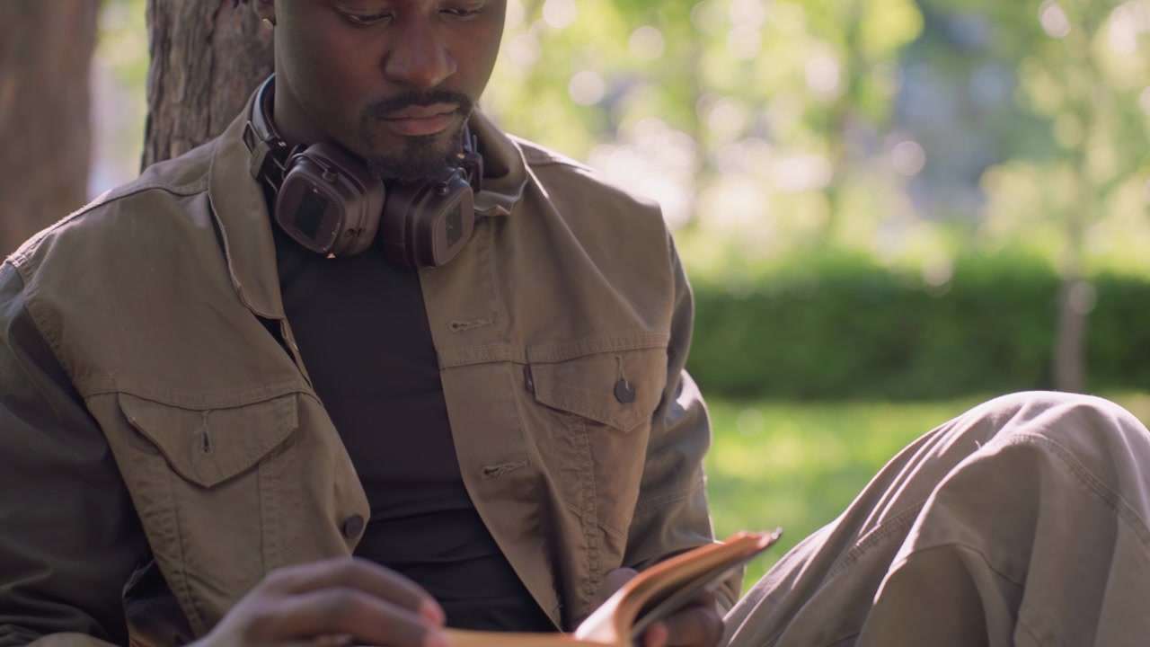 Primo piano di un uomo che legge un libro all'aperto in un parco, con le cuffie al collo. L'uomo indossa una giacca marrone e siede su una panchina sotto un albero, tenendo un libro aperto con le pagine visibili. Lo sfondo mostra...