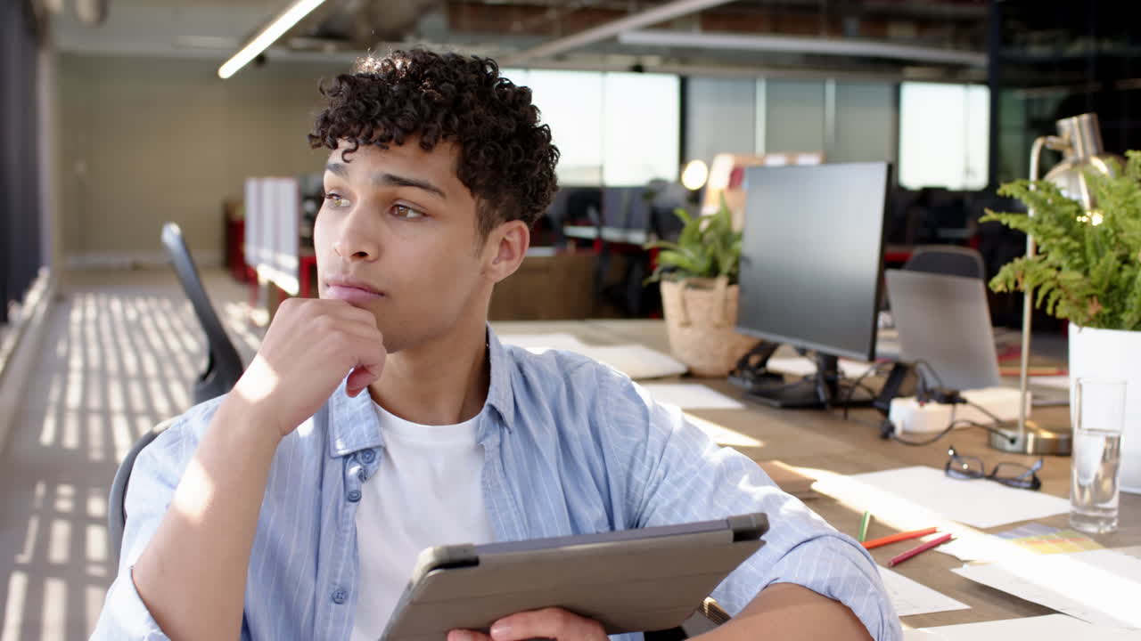 Holding tablet, man thinking while sitting at desk in modern office, copy space