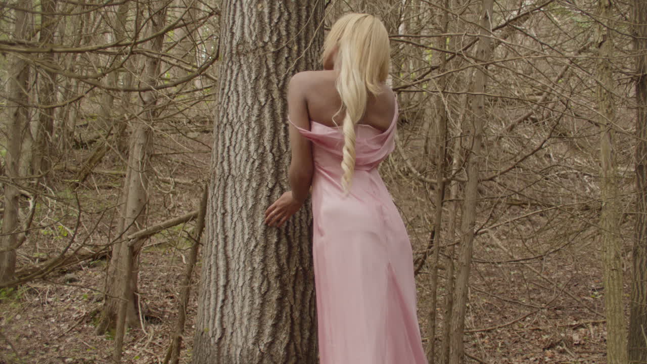 Young ebony woman, with long blonde hair, wearing a pink gown. She leans against a sweetgum tree in the forest, facing away from camera. She looks over shoulder towards camera, and then looks away.
