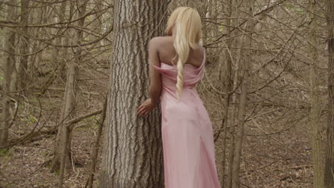joven mujer de ébano, con el cabello largo y rubio, con un vestido rosa. se apoya en un árbol de goma de azúcar en el bosque, mirando hacia lejos de la cámara. mira por encima del hombro hacia la cámara, y luego mira hacia lejos.