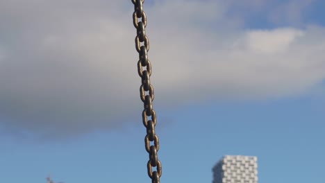 revealing close-up of yellow metal industrial chain and hook to lift cargo anchor and secure boats in the marina with a blurry cityscape in the background blue sky clouds buildings boats