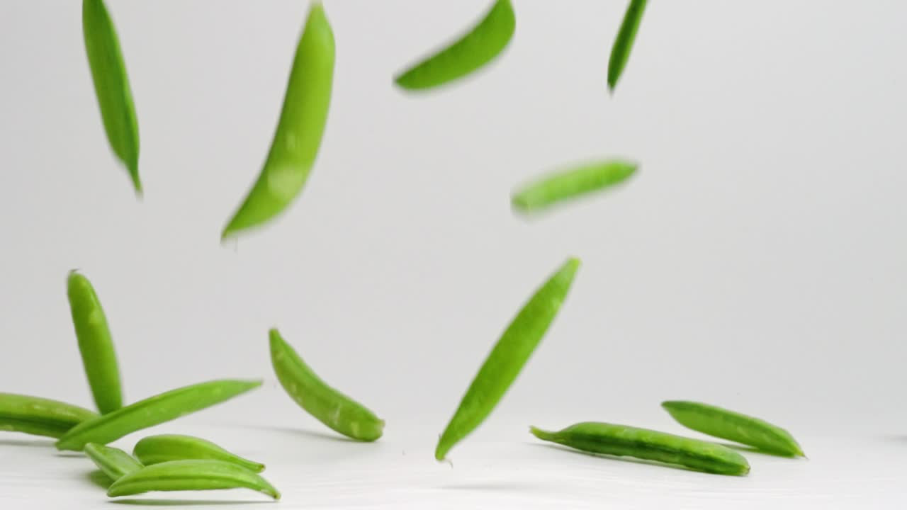 vainas de guisantes de azúcar enteras verdes frescas cayendo y rebotando en una pila sobre una mesa blanca en cámara lenta