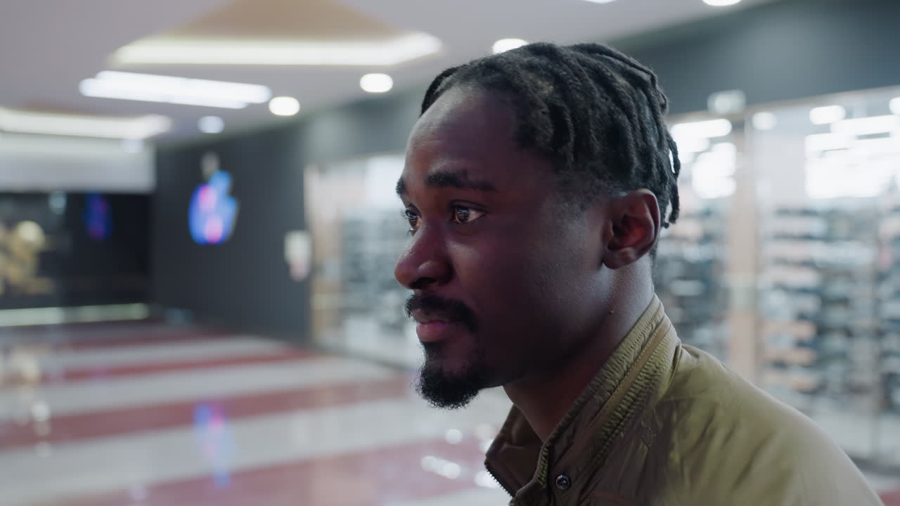 Music artist with braided hair wearing casual jacket looking contemplative in shopping mall with soft bokeh lights and blurred background expressing deep emotion and thoughtful mood