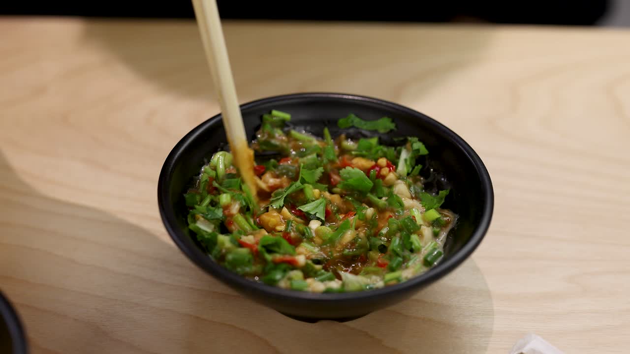 Hand stirs vibrant chili, garlic, and herb sauce in black bowl under soft indoor lighting