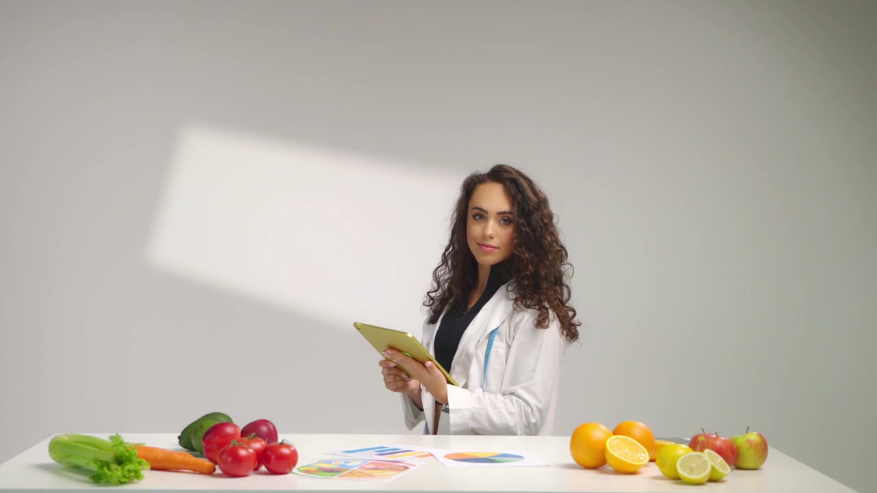 Dietologa in camice bianco seduta alla scrivania che esamina grafici nutrizionali colorati, circondata da frutta e verdura su entrambi i lati. Concetto di analisi alimentare sana e consulenza per una dieta equilibrata