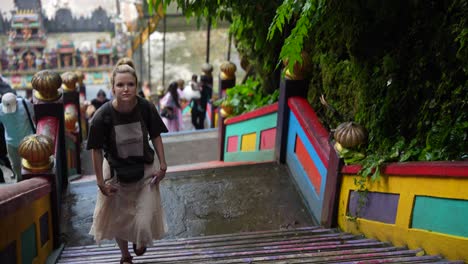 se ve a una mujer subiendo los coloridos escalones en la entrada de las cuevas batu en kuala lumpur, rodeada de barandillas vibrantes y vegetación exuberante, ubicación tropical