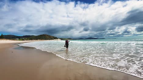 backpacker wades through surf at beach on cloudy day, slow motion wide shot