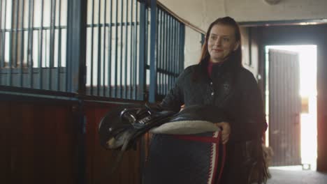 una mujer camina en el establo llevando una silla de caballo.