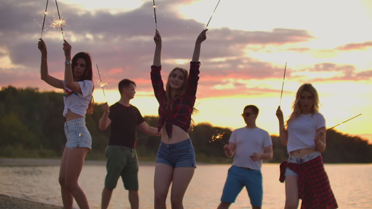 The young friends are dancing with big bengal lights on the sand coast. This is cool theamy summer evening on the open air party.