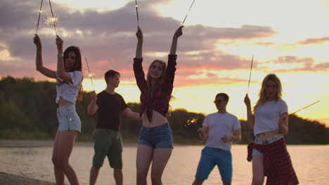 los jóvenes amigos están bailando con grandes luces bengalíes en la costa de arena. esta es una noche de verano genial en la fiesta al aire libre.