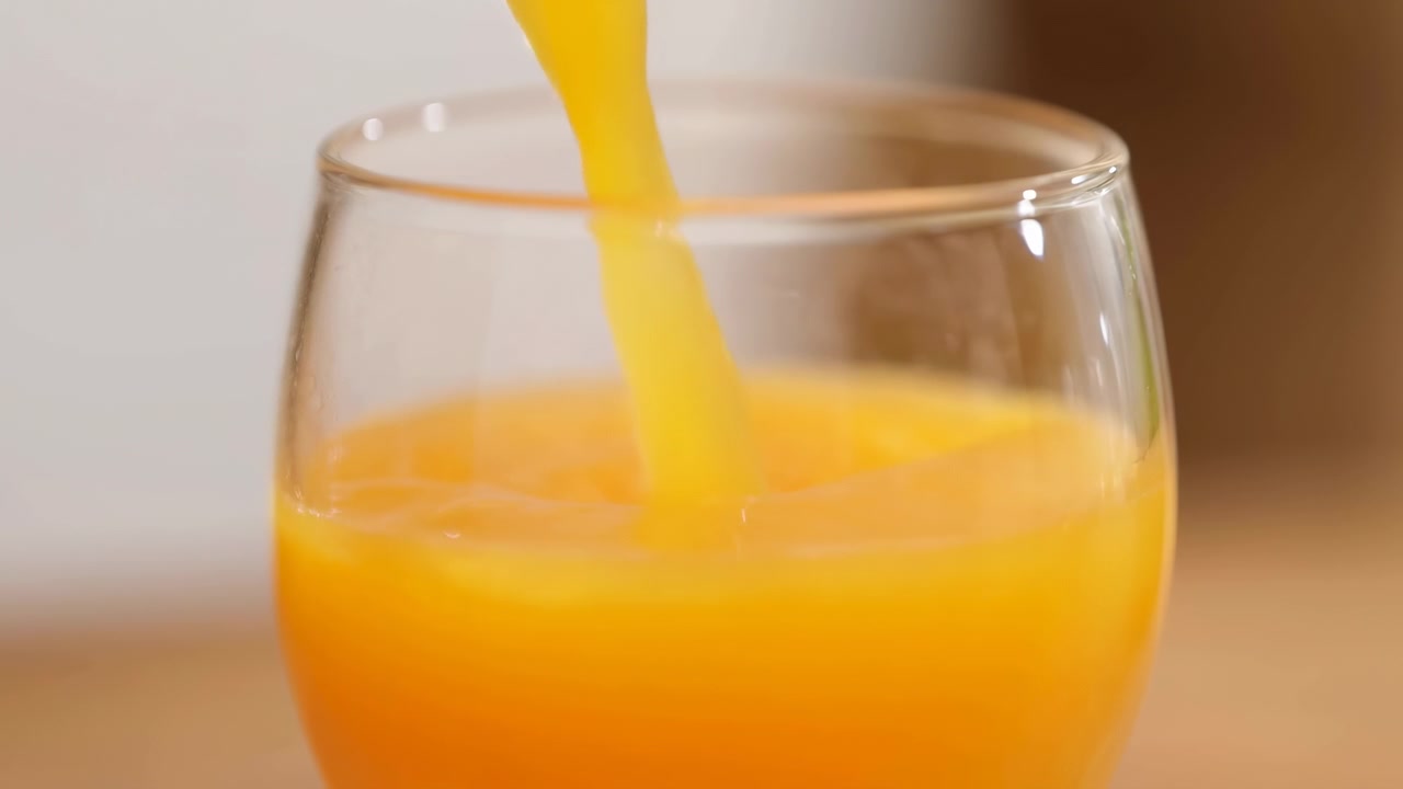 Klar appelsinjuice hældes i en jævn strøm ned i et klart stilkløst glas, hvilket hæver væskeniveauet, mens blød hvirvel, små bobler og blanke refleksioner skaber en frisk morgenmadspræsentation