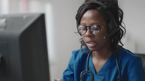 mujer negra médico usar bata blanca gafas y auriculares hablando con el cliente uso de videollamada. debido al brote de la pandemia de coronavirus terapeuta trabajando de forma remota proporcionar ayuda a los pacientes de la clínica por videollamada