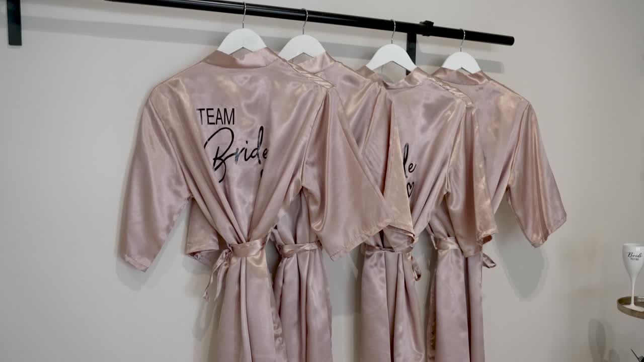 Quattro vestaglie abbinate sono appese in ordine su un appendiabiti. Ogni vestaglia riporta la scritta Team Bride. La stanza è luminosa e pronta per il giorno speciale che verrà. Le amiche si riuniscono per festeggiare insieme.