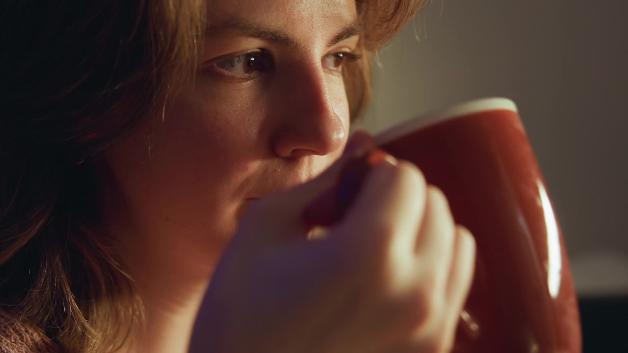 Close indoor view shows young woman holding hot ceramic mug, lifting it for gentle sips with relaxed posture, downward gaze, warm side lighting, cozy sweater, soft focus, peaceful home mood