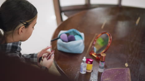 young girl in plaid shirt carefully holds and examines makeup items before placing them back in her bag, wooden table features beauty products, mirror, and makeup tools