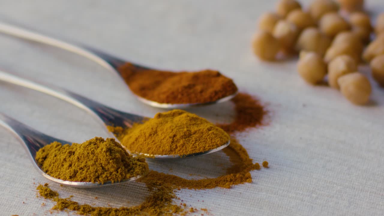 A spoonful of curry powder is placed on a table and some powder falls on the white tablecloth, nearby are two more spoons with turmeric powder and saffron powder and some chickpeas