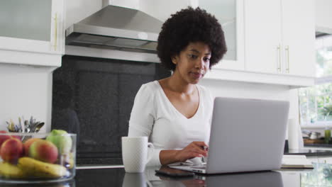 mujer afroamericana usando una computadora portátil en la cocina mientras trabaja desde casa