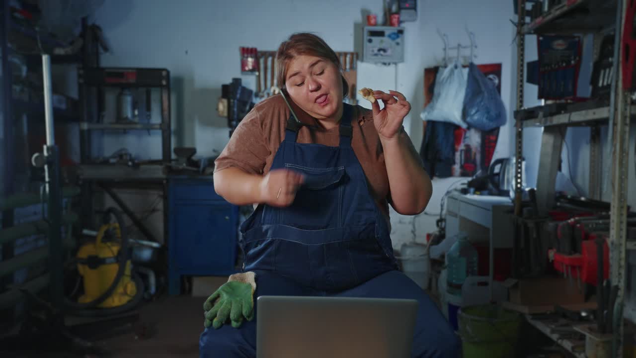 Woman Mechanic Eats Snack While Working on Laptop in Garage