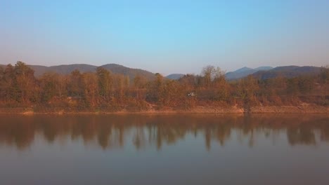 The-drone-shot-captures-a-car-driving-along-the-lakeshore,-set-against-a-backdrop-of-mountains-during-sunset