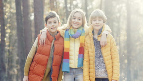 Close-up-view-of-joyful-kids,-girl-and-two-boys,-hugging-and-smiling-at-camera-in-the-forest-on-a-sunny-day