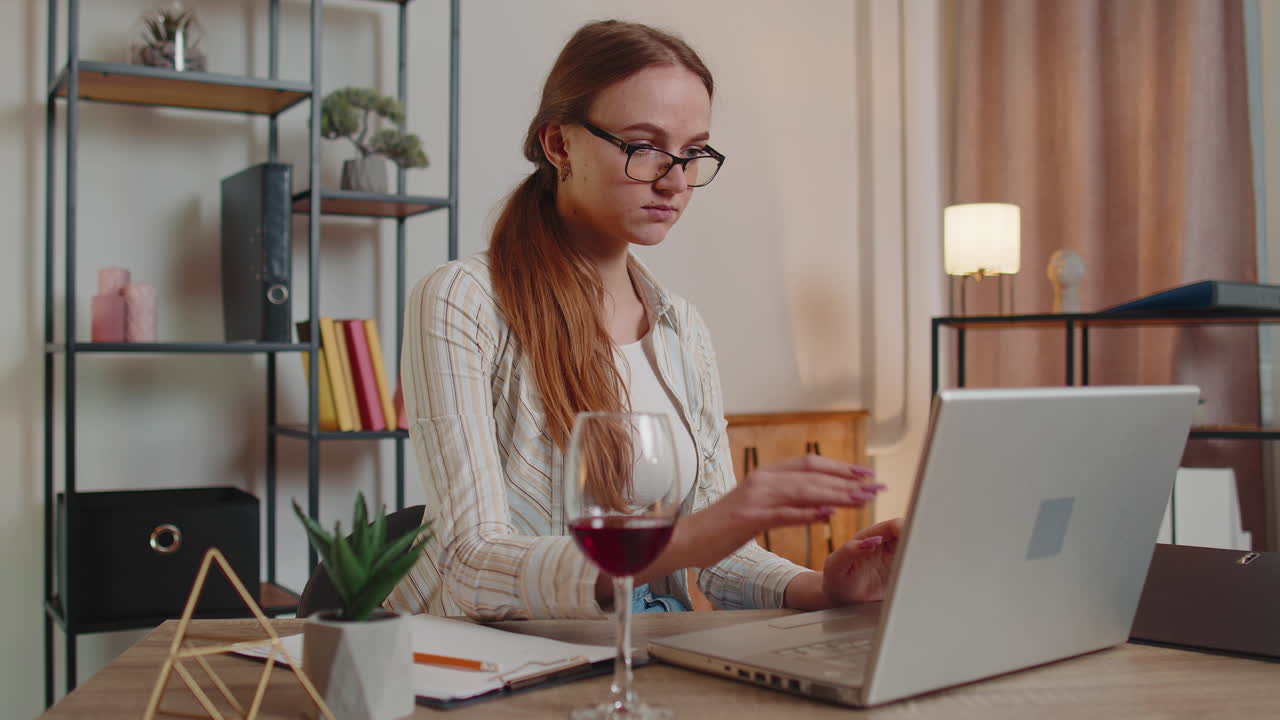 Eine konzentrierte freiberufliche Frau trinkt Wein, während sie zu Hause im Homeoffice online auf ihrem Laptop tippt