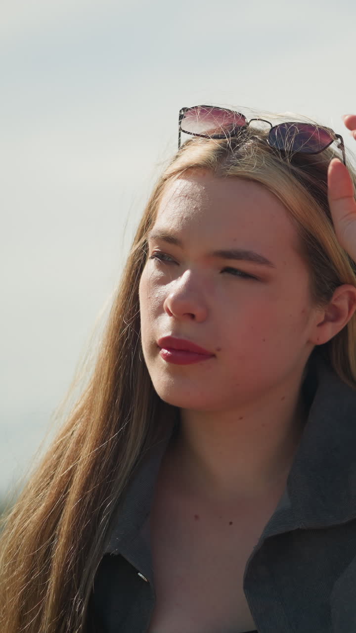 White lady adjusts sunglasses resting on her head with a focused expression, then adjusts her hair, with a blurred background featuring light poles in an outdoor urban setting on a sunny day