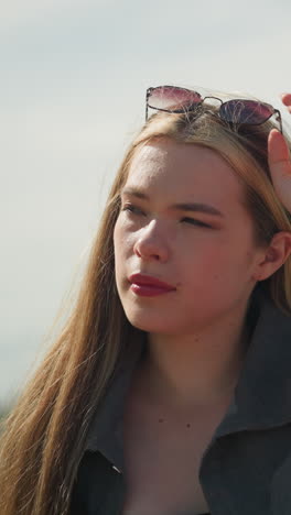dama blanca ajusta gafas de sol que descansan en su cabeza con una expresión enfocada, luego ajusta su cabello, con un fondo borroso con postes de luz en un entorno urbano al aire libre en un día soleado