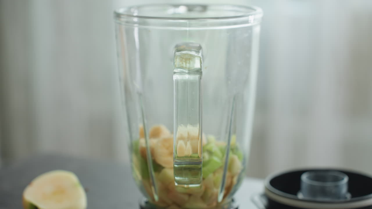 Forberedelse av gravid-smoothie: nærbilde av banan- og avokado-blender, klar glasskrukke, hånd som slipper skivede banan- og grønne avokadobiter på benkeplaten, lokk ved siden av krukken, mykt naturlig lys, morgenkjøkkenforberedelse.