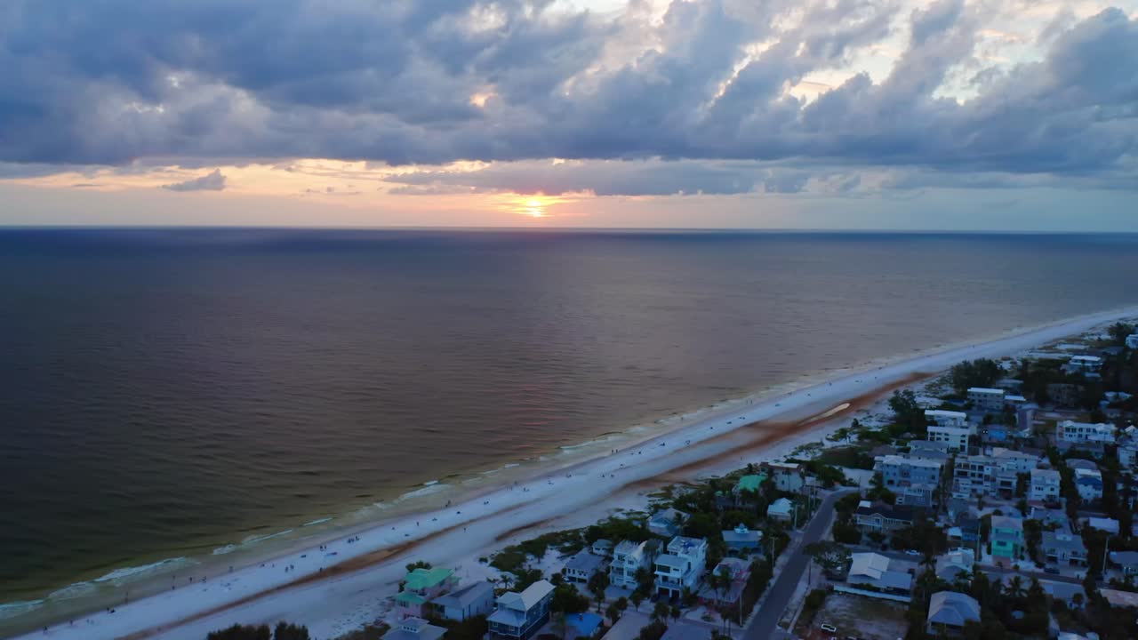 The last light of day glows over Anna Maria Island, illuminating calm Gulf waters and the white sandy shoreline as clouds drift above seaside homes in tranquil twilight