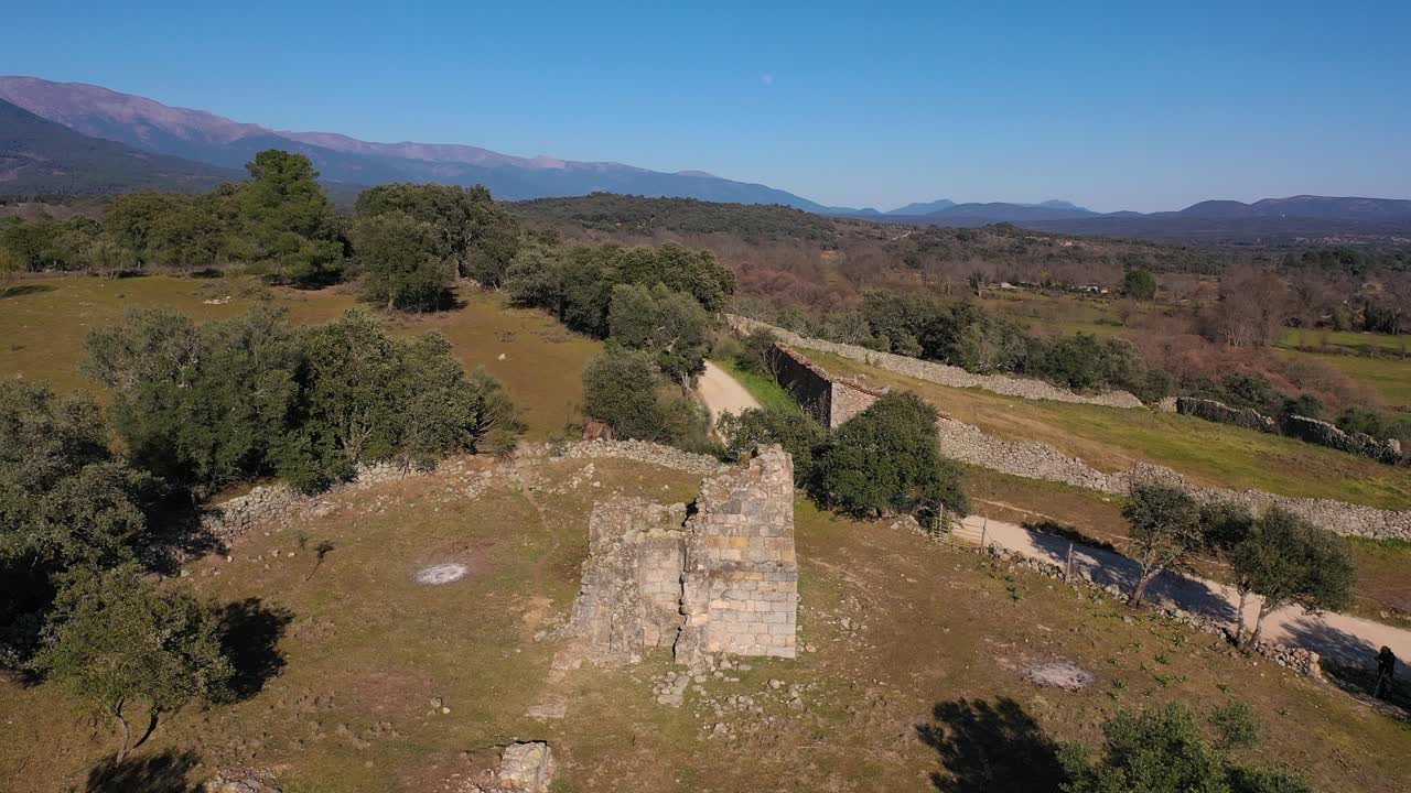 vuelo de avión no tripulado en reversa con una ermita medieval en ruinas del siglo xi rodeada de campos con pastos y bosques, en el fondo las montañas de la sierra de gredos en ávila, españa