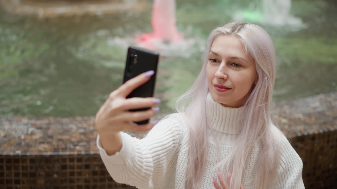 High angle view of stylish trendsetter seated behind mall water fountain adjusting her hair and smiling while taking selfie on mobile phone against softly blurred fountain backdrop bustling shopping