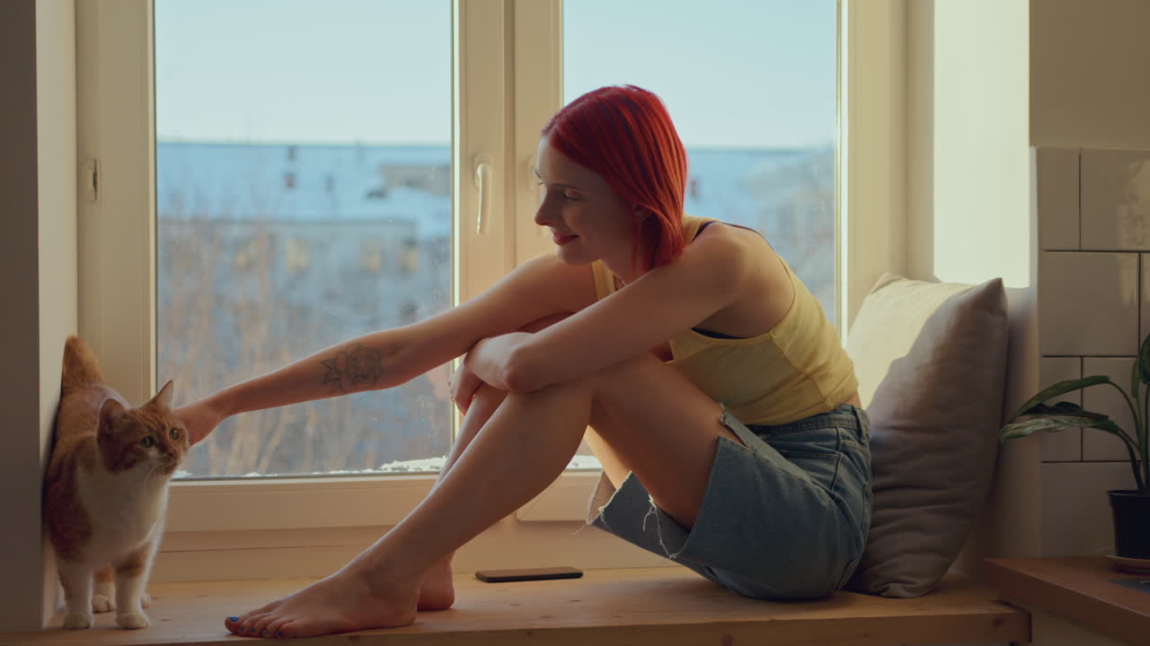 젊은 여성이 미소 짓고 집에서 창문 앞에 있는 고양이를 부드럽게 만지고 있다