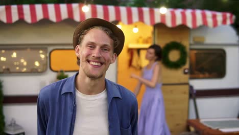 Happy-Cheerful-Couple-Portrait-Of-A-Man-In-Hat-Smiling-At-Foreground,-Woman-Having-Fun-Time,-Dancing-Together-On-Doorstep-House-On-Wheels-Outdoors-On-Blurred-Background