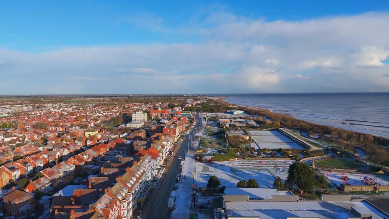 Una vista aerea mostra una città costiera innevata. Edifici con tetti rossi fiancheggiano le strade. Le strade sul lungomare mostrano la quiete invernale. Cielo nuvoloso sopra la scena. Nessuna persona presente