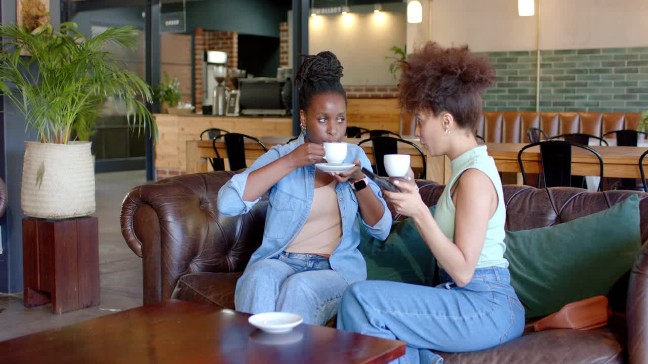 Afroamerikanische Freunde treffen sich im Café, sitzen auf dem Sofa mit weißen Tassen, überprüfen ihre Handys und unterhalten sich.