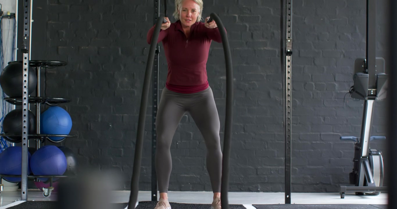 Donna di mezza età in palestra, sul tappetino, in felpa, che afferra le battle ropes e aumenta le onde per il fitness.