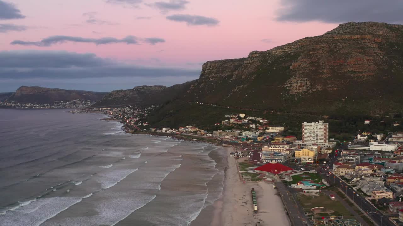 Aerial drone view captures city buildings nestled between majestic arid mountain and beach during sunset in Iceland. The warm golden light of setting sun bathes landscape on coast