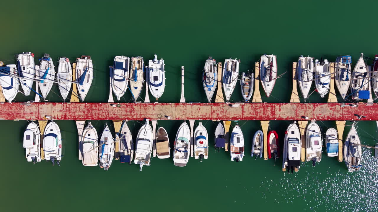 Vista dall'alto di numerose barche bianche e barche a vela ormeggiate lungo un molo di legno rosso circondate da acqua verde in un porto turistico