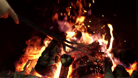 closeup on fire-iron placing burning logs in open indoor fireplace, flames on black background