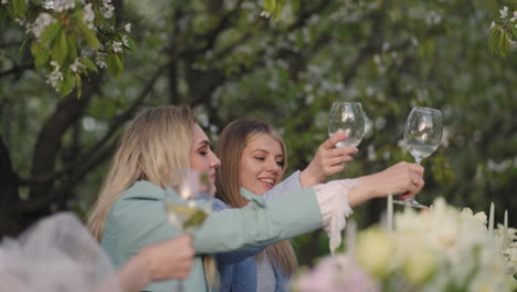 Alegre-Despedida-De-Soltera-En-Un-Jardín-Floreciente,-Las-Mujeres-Tintinean-Vasos-Y-Se-Ríen-Sentadas-En-La-Mesa-En-El-Huerto