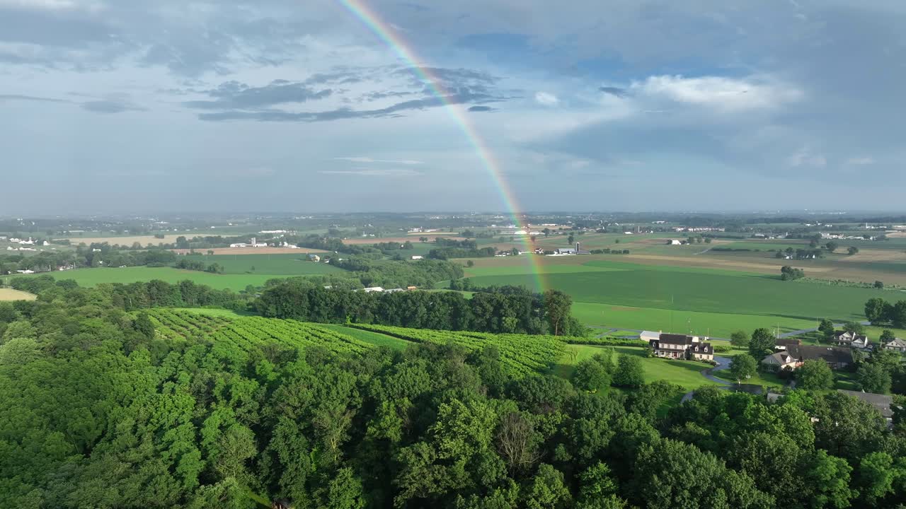 Aerial view of rural Pennsylvania, showing lush green fields and rolling hills under vast sky. Vibrant rainbow arches over landscape, adding touch of magic to serene countryside scene. Wide shot.