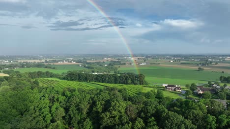 aerial view of rural pennsylvania, showing lush green fields and rolling hills under vast sky. vibrant rainbow arches over landscape, adding touch of magic to serene countryside scene. wide shot.