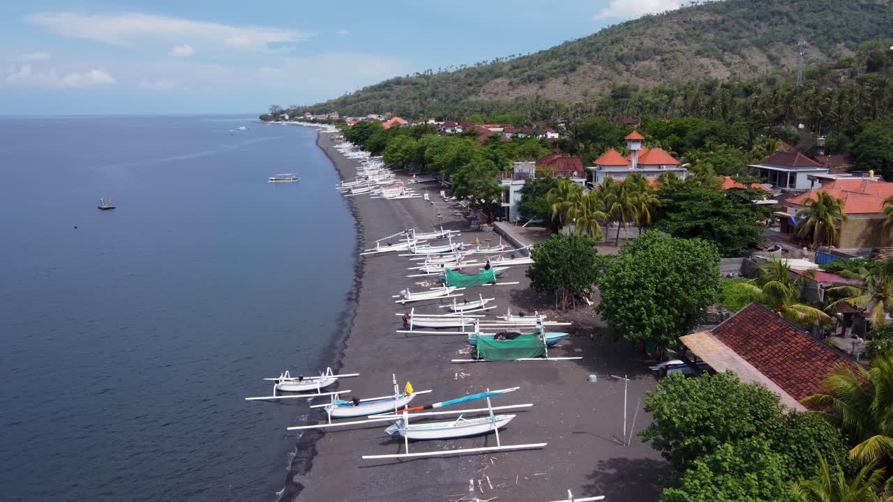 Aerial view of amed village seaside with local white Jukung fishing boats on black volcanic sand beach, villager houses and mountains in the background