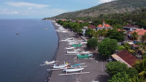 aerial view of amed village seaside with local white jukung fishing boats on black volcanic sand beach, villager houses and mountains in the background