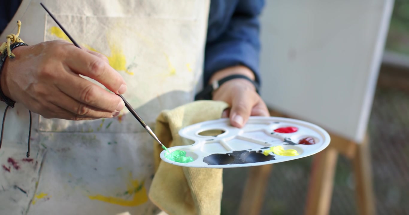 Anziana asiatica che intinge il pennello in un pozzetto verde sulla tavolozza, mescolando la pittura per la tela su un cavalletto all'aperto.