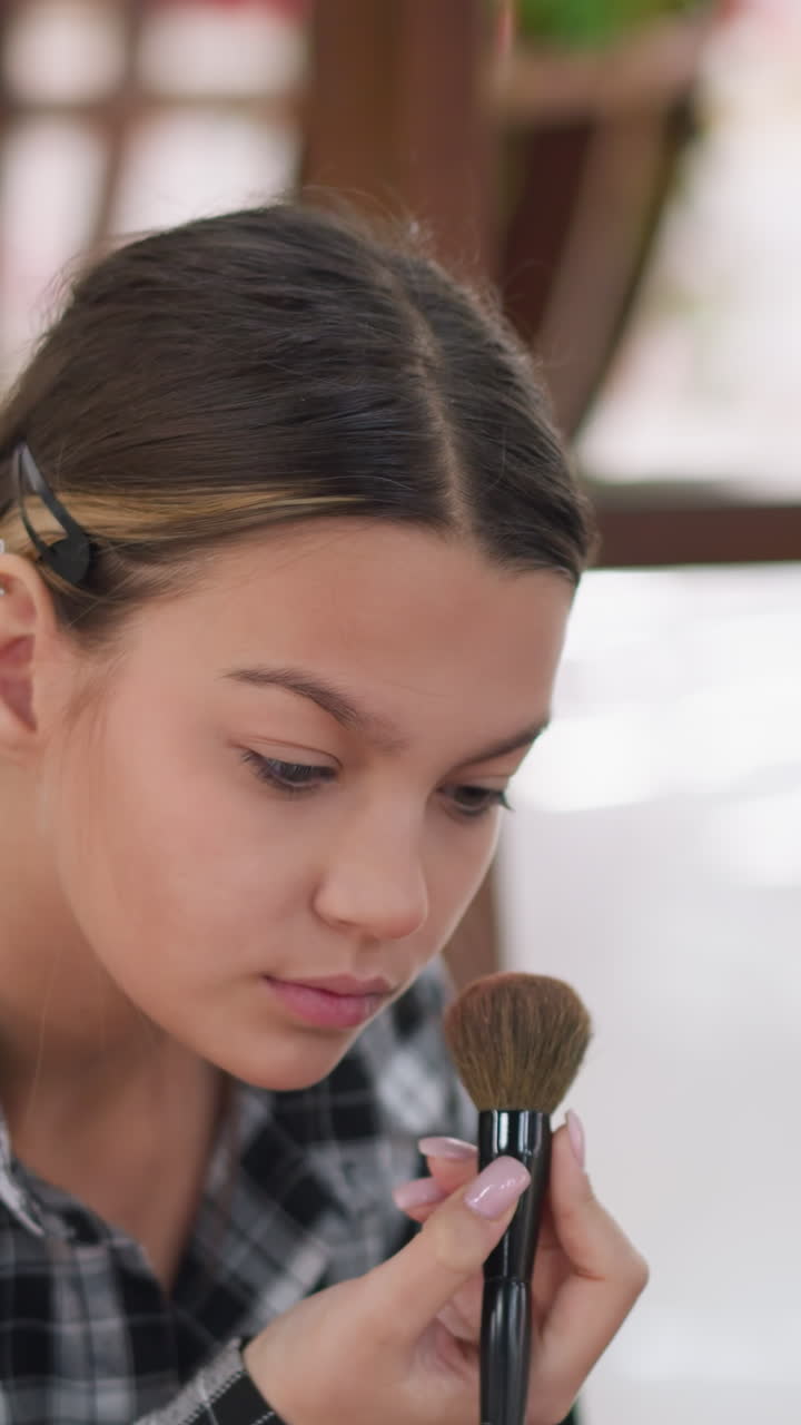Primer plano de adolescente concentrada practicando maquillaje con una brocha, con la brocha difuminando polvos e iluminador, borde del espejo visible, expresión atenta y mano firme, perfeccionamiento paso a paso de la técnica, uñas cuidadas